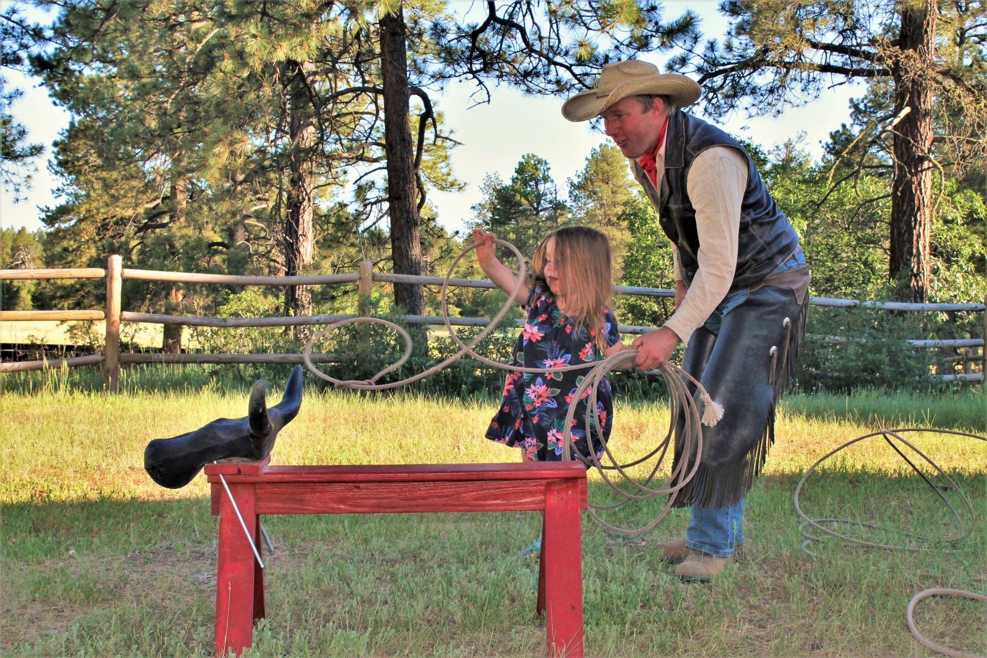 A little girl throws a rope at a pretend cow with a cowboy helping her